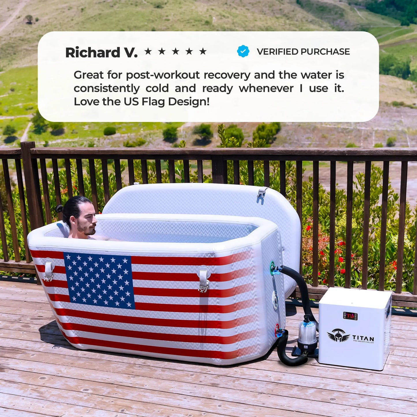 A man relaxes in a white rectangular Titan Wellness Triumph cold plunge tub. The front of the tub features a large American flag design. The tub sits on an outdoor wooden deck with a scenic mountain view. A white Titan Pro Water Chiller connects to the right side. The white tub lid rests open in the background. A text overlay displays a five star verified review from Richard V. The review praises the consistent cold water and the flag design.