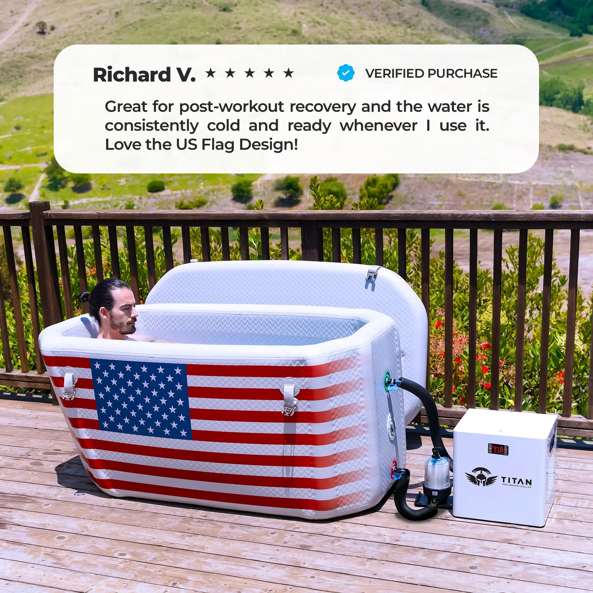 A man relaxes in a white rectangular Titan Wellness Triumph cold plunge tub. The front of the tub features a large American flag design. The tub sits on an outdoor wooden deck with a scenic mountain view. A white Titan Pro Water Chiller connects to the right side. The white tub lid rests open in the background. A text overlay displays a five star verified review from Richard V. The review praises the consistent cold water and the flag design.