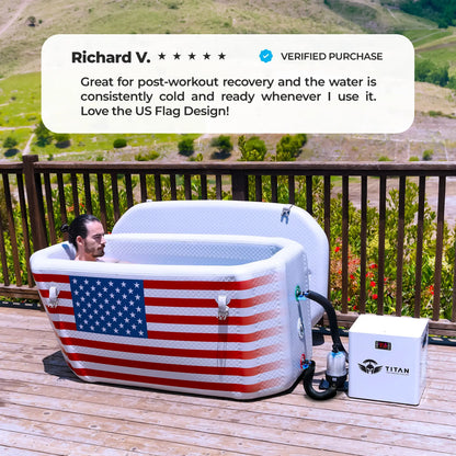 A man relaxes in a white rectangular Titan Wellness Triumph cold plunge tub. The front of the tub features a large American flag design. The tub sits on an outdoor wooden deck with a scenic mountain view. A white Titan Pro Water Chiller connects to the right side. The white tub lid rests open in the background. A text overlay displays a five star verified review from Richard V. The review praises the consistent cold water and the flag design.